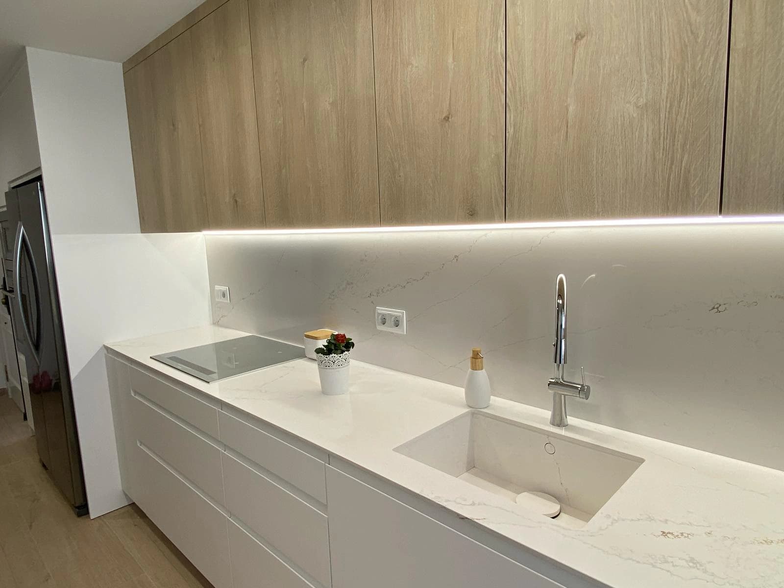 Vista general de una cocina blanca con encimera y fregadero integrados, frente de pared a juego y muebles altos de madera.